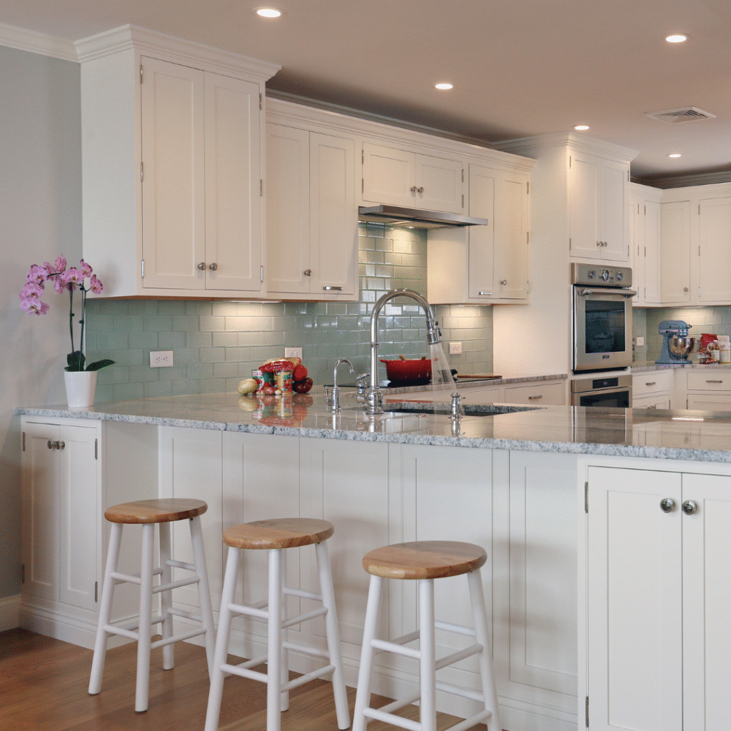 Bright and modern kitchen with white cabinetry, a marble island countertop, three wooden bar stools, and a chrome gooseneck faucet. Soft green subway tile backsplash and stainless steel appliances create a clean and elegant design.