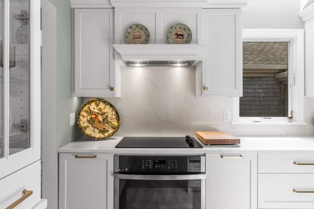 White kitchen with marble backsplash, centered range hood, and upper cabinets; decorative plates on the shelf above the stove.